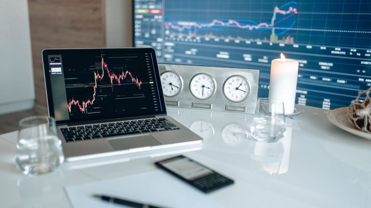 A laptop displaying trading charts on a desk with clocks and a candle in the background.