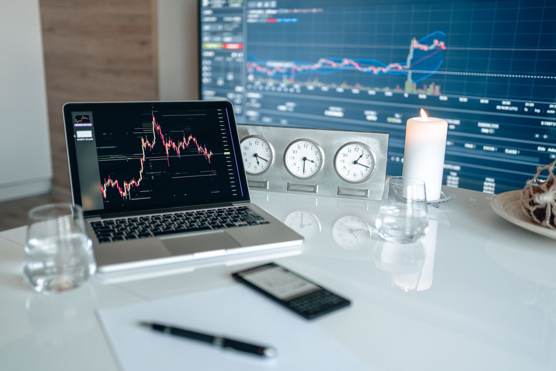 A laptop displaying trading charts on a desk with clocks and a candle in the background.
