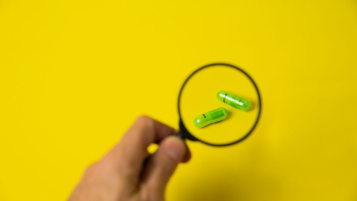 From above of crop unrecognizable male holding magnifying glass over green pills on yellow background in studio — Dr. Andrew