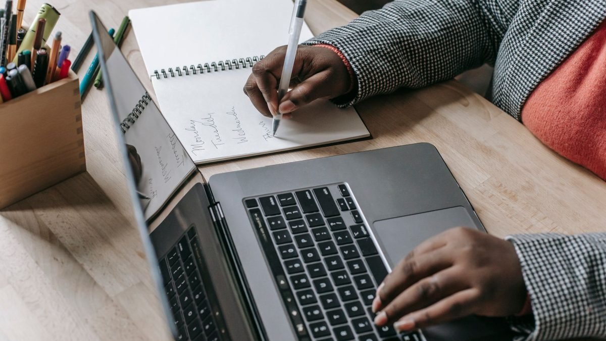 Crop unrecognizable African American female in formal wear writing notes in notepad while working on laptop — Enjoy Full-Bodi
